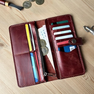 Könnte beinhalten: Offenes, weinrotes Lederportemonnaie mit mehreren Kartenfächern, einem Reißverschlussfach und einem Druckknopfverschluss. Das Portemonnaie ist mit Karten, Bargeld und Münzen gefüllt. Auf einer Holzoberfläche.