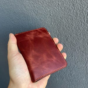 Puede incluir: Una cartera rectangular de cuero de color marrón rojizo oscuro con una textura suave ligeramente desgastada y costuras visibles a lo largo de los bordes. La cartera tiene un diseño plegado con una apariencia limpia y un tono de color rico y profundo.