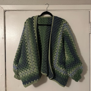 May include: A handmade, open-front crochet cardigan in shades of green and blue. The cardigan features a granny square pattern and long, voluminous sleeves. The garment is hanging on a black hanger against a white door.