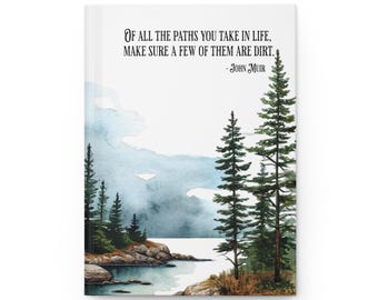 May include: A hardcover journal featuring a watercolor landscape of a lake, rocky shoreline, and evergreen trees. The text reads: Of all the paths you take in life, make sure a few of them are dirt. - John Muir. The cover has a white background.