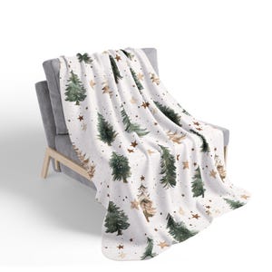 May include: A white throw blanket featuring a repeating pattern of watercolor-style dark green pine trees and small brown stars. The blanket is draped over a gray upholstered chair with light-colored wooden legs, showcasing the fabric texture.