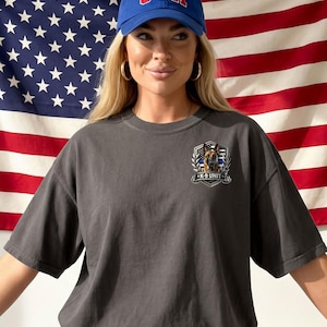 May include: A charcoal gray t-shirt featuring a K-9 Unit emblem with a German Shepherd dog, a blue line, and the text "K-9 UNIT." The model is wearing a blue baseball cap with "USA" in red letters. The American flag is in the background.