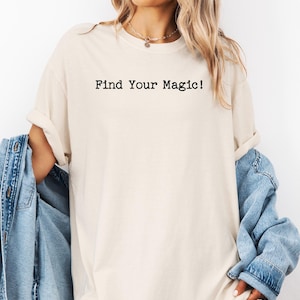 以下が含まれることがあります： 「Find Your Magic!」の黒い文字がプリントされたクリーム色のTシャツ。袖は折り返され、ライトブルーのデニムジャケットが羽織られています。全体的にカジュアルでリラックスした印象です。