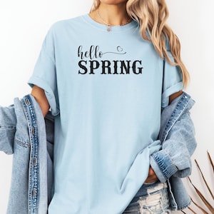 May include: Pale blue t-shirt featuring the text "hello SPRING" in black. The shirt has a casual fit and short sleeves. A denim jacket is worn over the shirt.