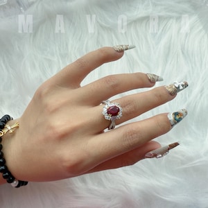 May include: A silver ring featuring a large oval ruby gemstone, encircled by smaller clear stones. The ring is displayed on a hand with long, decorated nails. A black beaded bracelet with a gold cross charm is also visible.