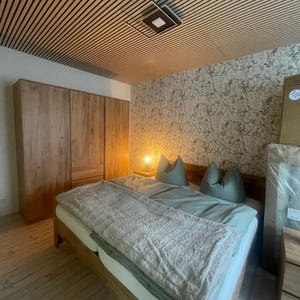 Puede incluir: Dormitorio con estructura de cama de madera, edredón y almohadas verde claro. Un armario de madera está a la izquierda y el techo tiene listones de madera. Una pequeña lámpara está sobre una mesita de noche.