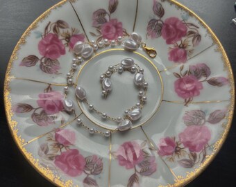 May include: A delicate gold chain necklace with white pearl accents. The necklace is displayed on a white porcelain plate with a floral pattern.