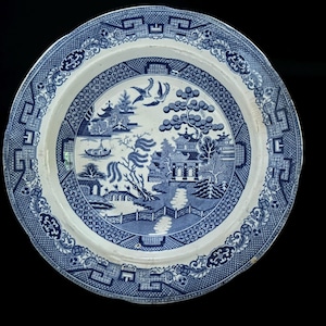 Puede incluir: Un plato de cerámica azul y blanco con un diseño detallado de patrón Willow. El plato presenta una escena central de paisaje con pagodas, árboles y un puente, rodeado por un borde decorativo. El plato es redondo y tiene un borde festoneado.