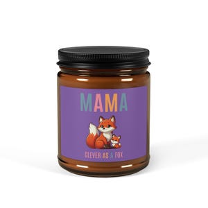 Op de afbeelding: Een paarse kaars pot met een bruin deksel. Het etiket zegt "MAMA CLEVER AS A FOX" in kleurrijke letters en heeft een cartoon illustratie van een rode vos en een vosje.