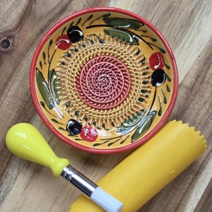 May include: A round ceramic grater plate with a yellow base, red rim, and hand-painted olive motifs in black, green, and red. The center features a raised spiral grating surface. Included is a yellow silicone garlic peeler and a small cleaning brush.