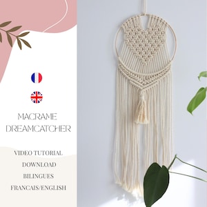 Op de afbeelding: Een witte macramé droomvanger met een hartvormig ontwerp en een gouden hoepel. De droomvanger hangt aan een witte muur en heeft een kwast aan de onderkant. De tekst "MACRAME DREAMCATCHER" is in zwarte letters op een roze achtergrond geschreven. De tekst "VIDEO TUTORIAL DOWNLOAD BILINGUES FRANCAIS/ENGLISH" is in zwarte letters op een witte achtergrond geschreven.