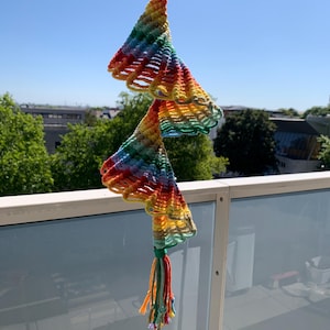 May include: A handmade wind chime with a rainbow-colored, crocheted design. The chime features a wooden ring at the top, two bell-shaped sections, and a tassel with a small brass bell at the bottom. The colors include red, orange, yellow, green, and blue.