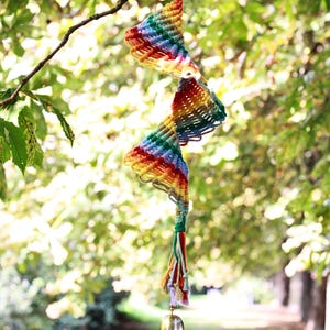 May include: A rainbow-colored macrame spiral hanging decoration with a small brass bell at the bottom. The spiral is suspended from a wooden ring and hangs against a blurred background of green foliage.