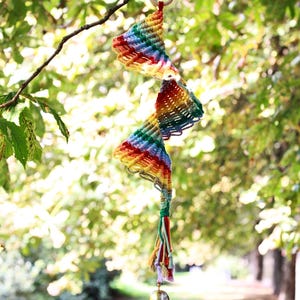 May include: A colorful macrame wind chime hangs from a tree branch. The chime features a spiral design with rainbow hues and a small bell at the bottom. The background is a blurred view of a green park.
