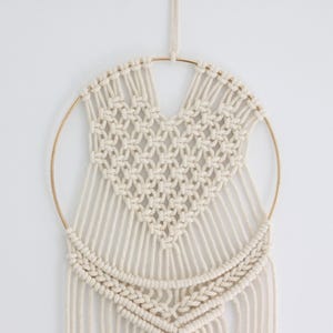 K&ouml;nnte beinhalten: Ein wei&szlig;er Makramee-Wandbehang mit einem herzf&ouml;rmigen Design in der Mitte. Der Behang ist aus Baumwollschnur gefertigt und hat einen goldenen Metallring.