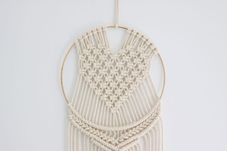 K&ouml;nnte beinhalten: Ein wei&szlig;er Makramee-Wandbehang mit einem herzf&ouml;rmigen Design in der Mitte. Der Behang ist aus Baumwollschnur gefertigt und hat einen goldenen Metallring.