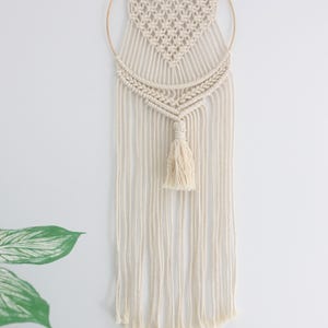 K&ouml;nnte beinhalten: Ein wei&szlig;er Makramee-Wandbehang mit Herzform und einem goldenen Ring. Der Behang hat unten einen Quasten.