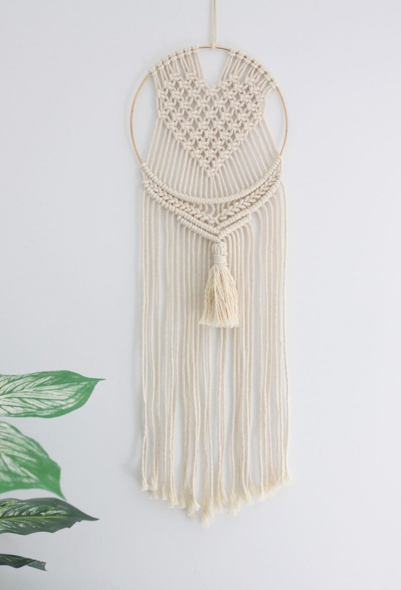 K&ouml;nnte beinhalten: Ein wei&szlig;er Makramee-Wandbehang mit Herzform und einem goldenen Ring. Der Behang hat unten einen Quasten.