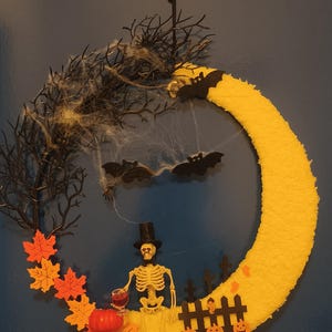 Puede incluir: Una corona de Halloween con forma de media luna amarilla, con un esqueleto con sombrero de copa que sostiene un vaso de líquido rojo. La corona incluye murciélagos negros, una calabaza y hojas naranjas y amarillas. El fondo es azul oscuro.