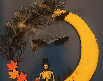 Może przedstawiać: Wieniec na Halloween w kształcie żółtego półksiężyca, ze szkieletem w cylindrze trzymającym kieliszek z czerwoną cieczą. Wieniec zawiera czarne nietoperze, dynię oraz pomarańczowe i żółte liście. Tło jest ciemnoniebieskie.