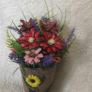 Könnte beinhalten: Dekorative Blumengesteck in einem mit Jute umwickelten Topf. Der Strauß zeigt leuchtend rote, rosa und lila Blumen mit grünen Akzenten. Eine gelbe Sonnenblume und weiße Gänseblümchen zieren die Außenseite des Topfes.