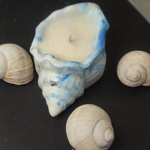 Op de afbeelding: Een schelpvormige kandelaar met een witte kaars en een lont. De houder is wit met blauwe accenten en zilveren vlekken. Drie schelpen zijn rond de kandelaar gerangschikt.