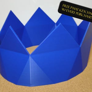 Pode incluir: Um objeto em forma de coroa geométrica azul vibrante. A coroa tem uma série de picos triangulares e uma base circular. O texto "FREE PHAT KEYCHAIN W/ EVERY PURCHASE!" é exibido em um retângulo preto.