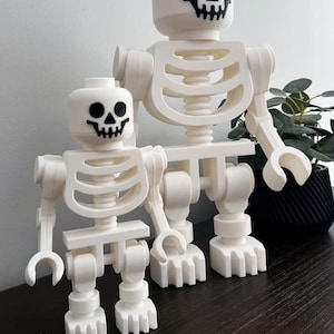 May include: Three white skeleton figures, resembling Lego toys, are displayed on a dark surface. Each figure has a black skull face. A small skeleton figure is in front of the larger ones. The figures are a Halloween or spooky theme.