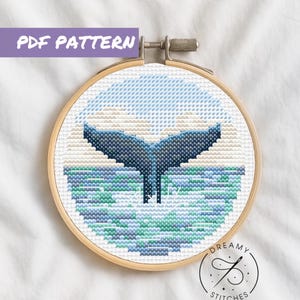 May include: A cross stitch pattern featuring a whale tail emerging from the ocean. The pattern is in a wooden embroidery hoop and includes a blue, white, and green colour scheme. The pattern is labelled "Dreamy Stitches" with a social media handle.