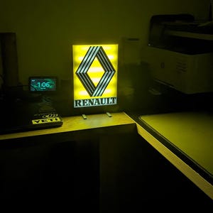 May include: A rectangular illuminated sign displaying the Renault logo, a diamond shape with black and silver stripes on a yellow background. The word "RENAULT" is below the logo in white. The sign is lit up.