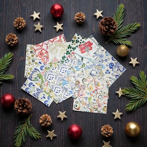 Può includere: Una collezione di tovaglioli di carta fantasia con disegni floreali e a spirale, disposti su una superficie di legno scuro. I tovaglioli sono circondati da decorazioni natalizie, pigne e decorazioni a stella dorata, creando un tema festivo.