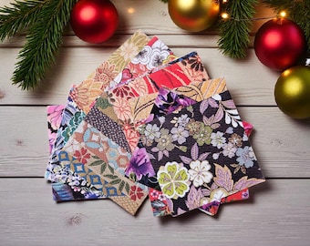 May include: A stack of square paper napkins featuring a variety of colourful floral and geometric patterns in shades of black, purple, red, gold, and blue, arranged in a fan shape on a light-coloured wooden surface with pine branches and ornaments.