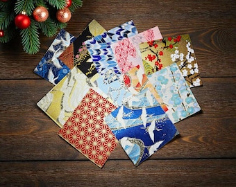 May include: A collection of square fabric coasters featuring various Japanese-inspired patterns, including white cranes, red and gold geometric shapes, blue floral motifs, and cherry blossoms, arranged in a fan-like display on a dark wood surface.