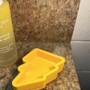以下が含まれることがあります： 黄色のプラスチック製のパイナップル型の皿。皿は茶色のカウンタートップの上に置かれています。ボトルのテキストは「Clean Skin Club Pineapple Glow Mist Hyaluronic Acid Peptides Antioxidants 100 ml / 3.4 fl. oz.」です。