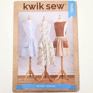 May include: A Kwik Sew pattern package showing three apron designs on dress forms. The aprons include a blue and white striped apron, a floral apron, and a brown apron. The package has the text "Kwik Sew" and "Misses' Aprons". The aprons are displayed on wooden stands.