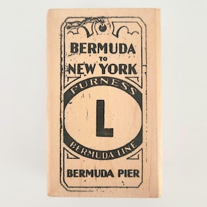 May include: A rectangular wooden block with black text that reads "BERMUDA TO NEW YORK FURNESS L BERMUDA LINE BERMUDA PIER". The text is inside a decorative frame with an oval shape in the center containing the letter "L".