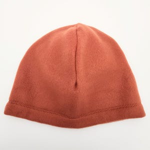 May include: A rust-colored beanie hat made of soft fleece material. The hat has a simple design with a stitched hem around the bottom. This warm hat is suitable for cold weather and outdoor activities.