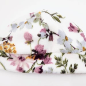 May include: A white beanie hat with a floral design. The hat has a soft, fuzzy texture and is decorated with a repeating pattern of purple, yellow, and blue flowers and green leaves.