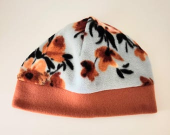 May include: A fleece beanie hat with a floral pattern. The hat features a light blue background with orange and black floral designs. The brim is a solid orange color.