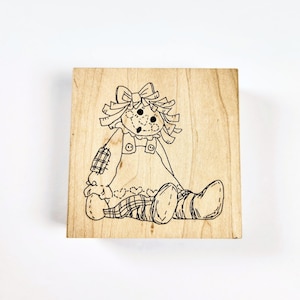 May include: A square wooden rubber stamp featuring a black line art illustration of a rag doll with yarn hair, a bow, a dress with buttons, and a patch on the sleeve. The doll is depicted in a seated position with legs extended forward.