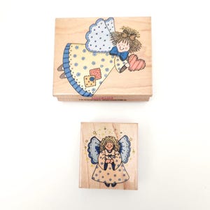 May include: Two wooden stamps with angel designs. The top stamp features an angel in a yellow dress with blue polka dots and a blue bow. The bottom stamp shows an angel in a similar dress with a smiling face and star accents.