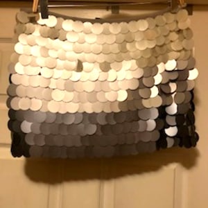 May include: A mini skirt made of overlapping, circular sequins in shades of white, gray, and black, hanging on a wooden hanger. The skirt's design creates a gradient effect, transitioning from light to dark.