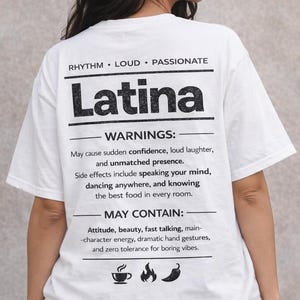 May include: White t-shirt with the word "Latina" in bold black letters. The shirt includes text about confidence, laughter, and attitude. The shirt has a crew neck and short sleeves.