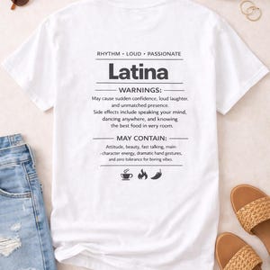 May include: White t-shirt with the word "Latina" printed in black, and a list of "warnings" and "may contain" attributes. The shirt is displayed with sunglasses, sandals, and denim shorts.
