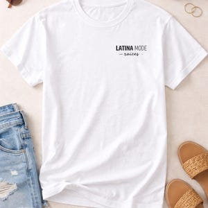 May include: A white t-shirt with the text "LATINA MODE - raices" printed in black. The shirt is displayed with denim shorts, sunglasses, sandals, a handbag, and jewellery.