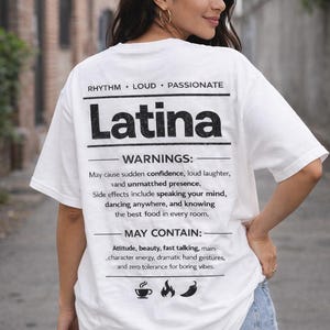 May include: White t-shirt with the word "Latina" in bold black letters. The shirt also features text that reads "Rhythm, Loud, Passionate" and a list of "Warnings" and "May Contain" statements. The model is wearing denim shorts.
