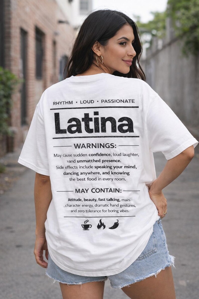 May include: White t-shirt with the word "Latina" in bold black letters. The shirt also features text that reads "Rhythm, Loud, Passionate" and a list of "Warnings" and "May Contain" statements. The model is wearing denim shorts.