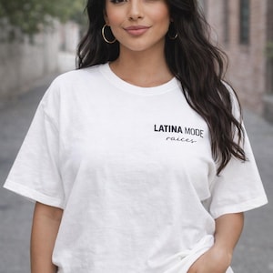 May include: White t-shirt with the text "LATINA MODE" and "raices" in black. The shirt is worn by a person with dark hair and gold hoop earrings. The person is wearing blue jeans and has their hand in their pocket.