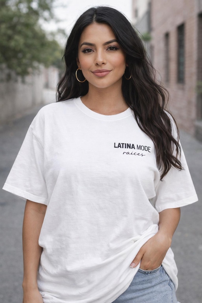 May include: White t-shirt with the text "LATINA MODE" and "raices" in black. The shirt is worn by a person with dark hair and gold hoop earrings. The person is wearing blue jeans and has their hand in their pocket.