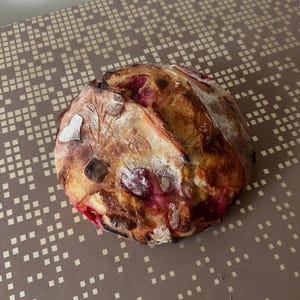 May include: A circular loaf of artisan bread with a golden-brown crust and visible red fruit inclusions. The bread has decorative leaf-like patterns and a dusting of flour on top, suggesting a rustic, homemade style.